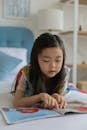 Adorable smart Asian girl reading magazine with bright illustrations while resting on bed