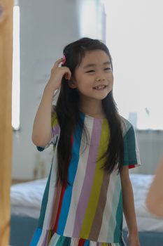 一位开朗可爱的亚洲女孩穿着明亮的家居礼服，对着镜子欣赏自己的发型