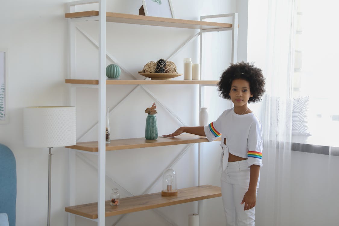 Serious little African American girl wiping dust from shelf of rack in room looking at camera