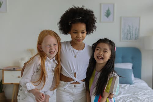Group of charming multiethnic girls embracing and laughing while standing against bed at home