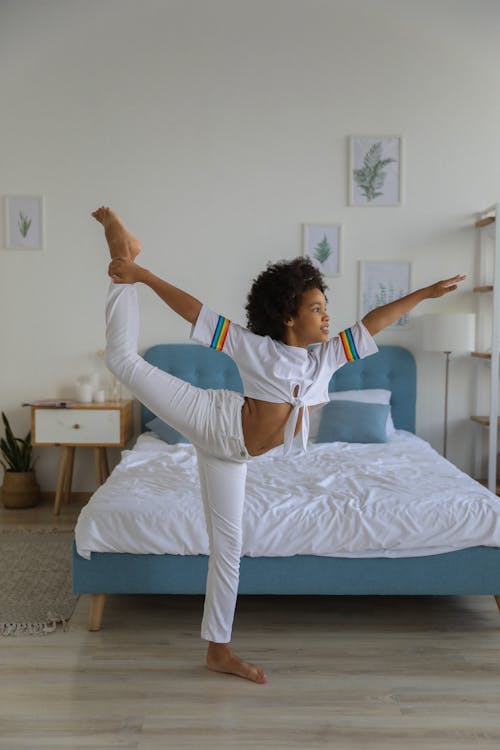 Free Full length of black girl in white clothes performing acrobatic splits standing against bed and looking forward Stock Photo