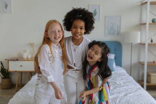 Group of multiethnic charming girls having fun and hugging while standing at bed in apartment