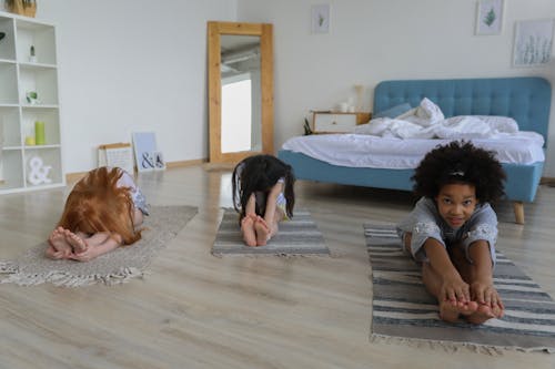 Little diverse girlfriends sitting on rugs and practicing Seated Forward Bend in light bedroom