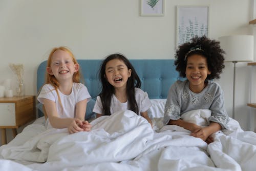 Laughing multiethnic best friends enjoying time during sleepover lounging under blanket in bed