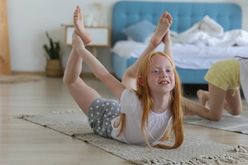 Free Full body of cheerful girl in casual clothing with reddish hair looking away with smile while stretching legs on carpet in bright cozy living room on blurred background Stock Photo