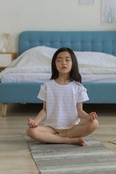 A young girl in a peaceful meditation pose on a mat indoors, embodying calm and mindfulness.