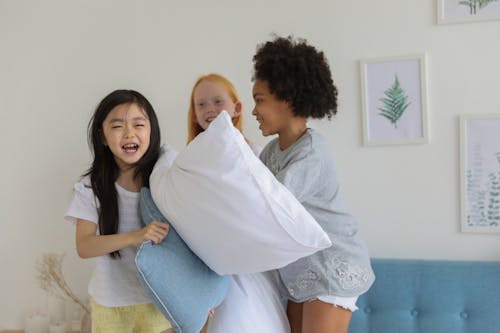 Happy diverse children having pillow fight while laughing cheerfully on bed in daytime