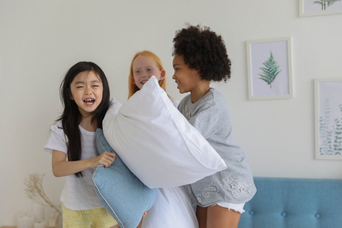 Happy diverse children having pillow fight while laughing cheerfully on bed in daytime