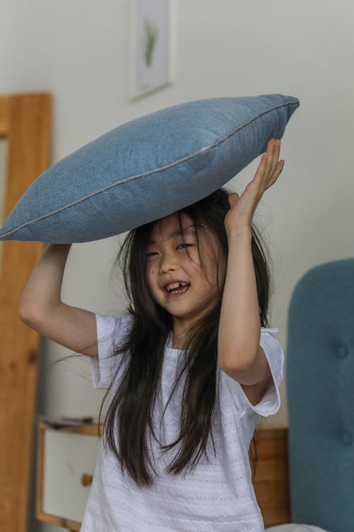 Cheerful Asian girl in casual clothing with long black hair looking at camera with bright smile while holding blue cushion over head in bright bedroom