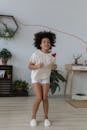 Full body of cheerful African American girl with curly hair looking away with bright smile while jumping over skipping rope in bright room