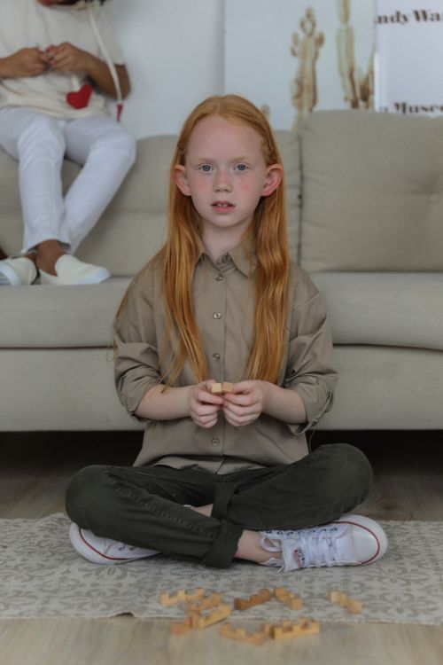 Free Full body of cute red haired child in stylish clothes sitting on floor and playing game while looking at camera Stock Photo