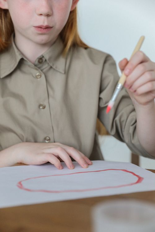 Free Crop unrecognizable little girl with foxy hair sitting at wooden table and painting with paintbrush and gouache on sheet of paper Stock Photo