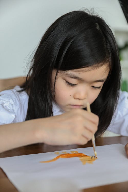 Cute attentive little ethnic schoolgirl drawing with paintbrush and watercolor paints while sitting at table during art lesson