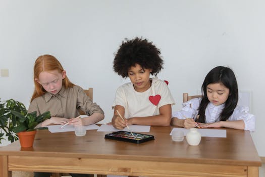 Group of adorable multiracial schoolgirls sitting at wooden table and drawing together with aquarelle during lesson