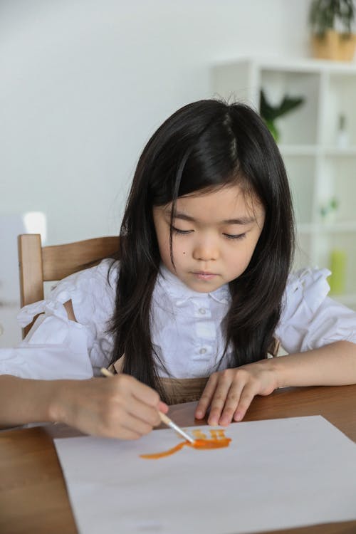 Free From above of concentrated little Asian schoolgirl in white shirt drawing on paper sheet with watercolor using paintbrush in classroom Stock Photo