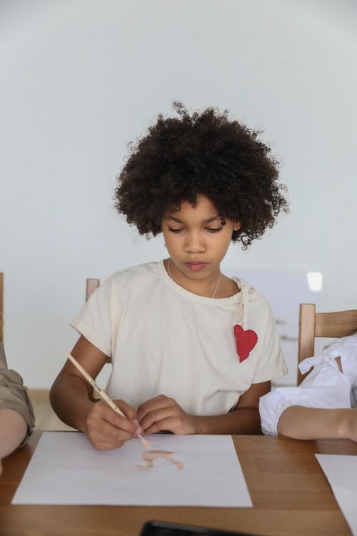Free Concentrated talented African American girl with curly hair using paintbrush while drawing with watercolor sitting at table with unrecognizable friends Stock Photo