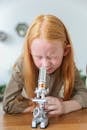 Curious little girl with long ginger hair sitting at wooden desk and examining samples under microscope in science lab