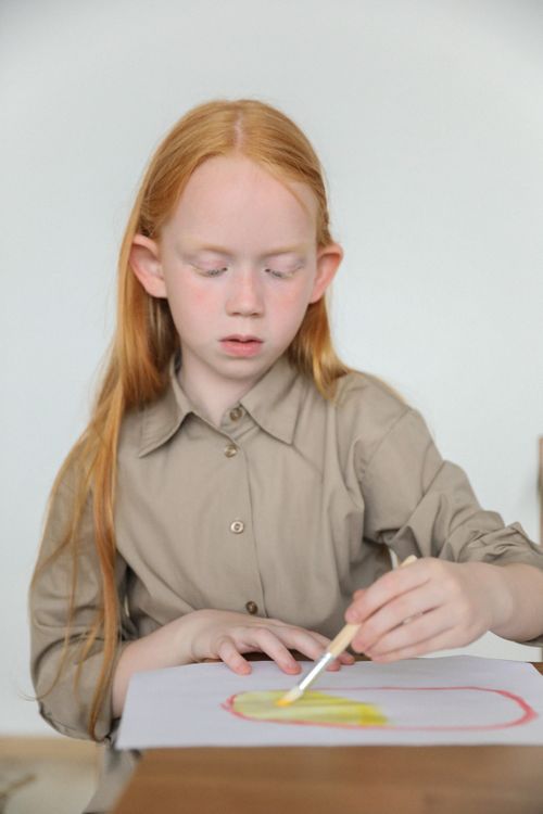 Free Adorable thoughtful preteen girl with long red hair sitting at table and painting on paper sheet using brush and watercolors Stock Photo