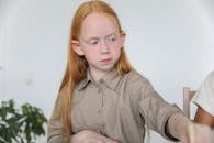 Attentive little girl with ling ginger hair in casual clothes dipping paintbrush into water while sitting at table during art lesson