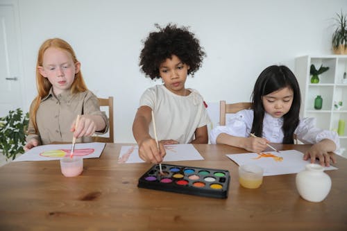 Talentierte Verschiedene Kleine Mädchen, Die Auf Papieren Mit Aquarellen Malen, Während Sie Zusammen Am Tisch Sitzen