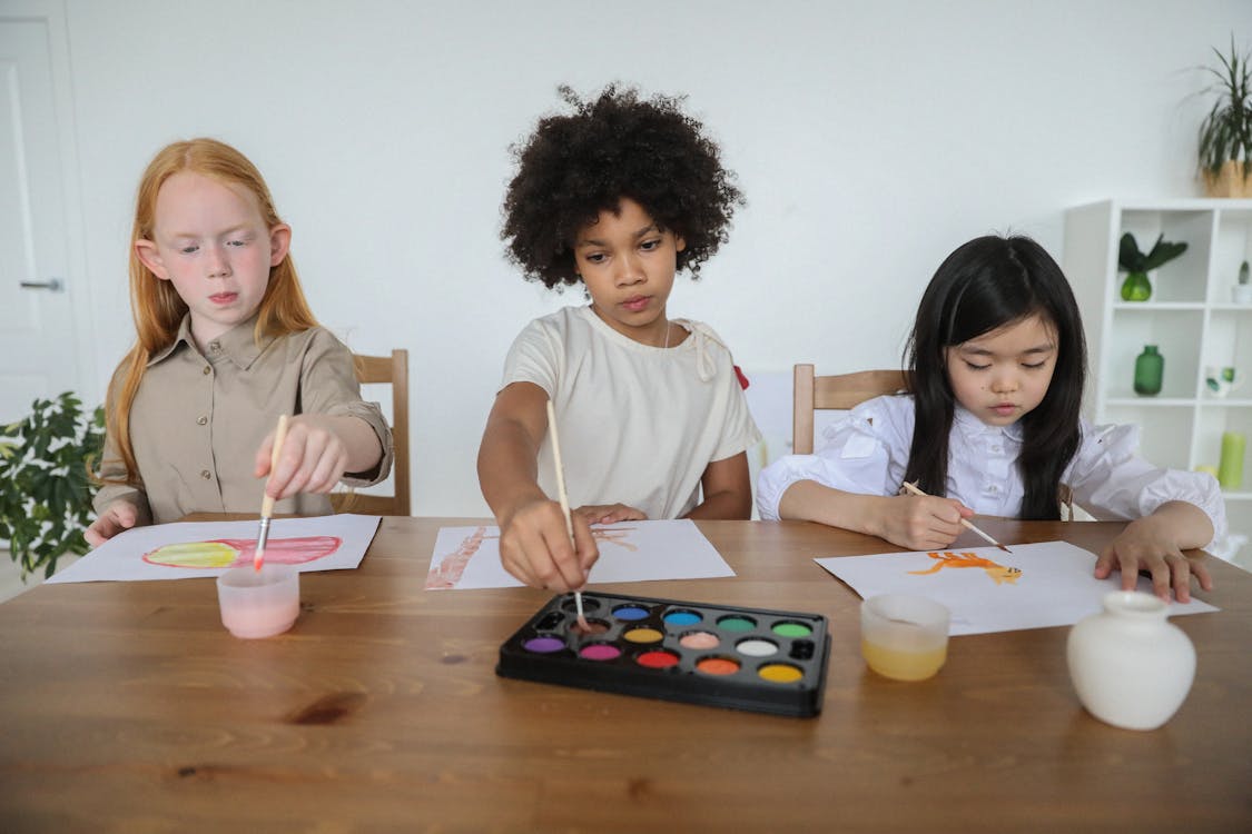 Free Talented diverse little girls painting on papers with watercolors while sitting together at table Stock Photo
