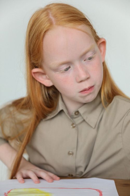 Free Concentrated little girl with long ginger hair sitting at table and looking away while painting with watercolors in classroom Stock Photo