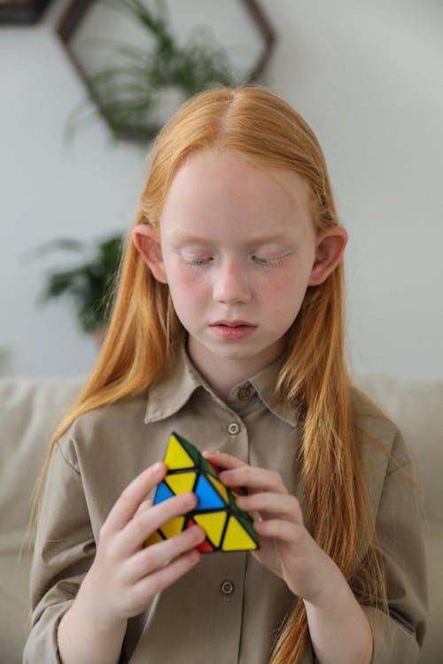 Smart concentrated little girl with long ginger hair playing with pyramid shaped 3D combination puzzle sitting on sofa at home