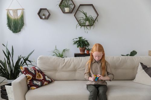 Free Cute smart girl solving bright puzzle while sitting on cozy sofa in comfortable living room decorated with green potted plants Stock Photo