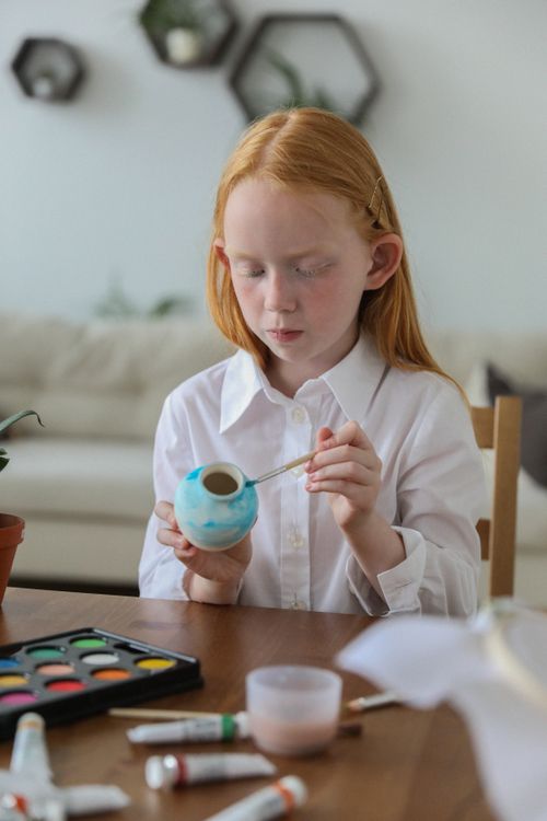 Free Cute small girl with red hair drawing on vase with watercolor while sitting at table Stock Photo