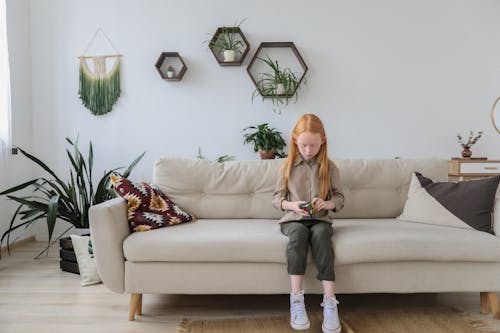 Serious girl in fashionable clothes solving puzzle and sitting on cozy sofa with bright cushion in contemporary lounge