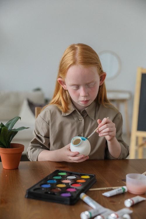 Free Concentrated creative adorable girl with red hair painting on vase with brush on table Stock Photo