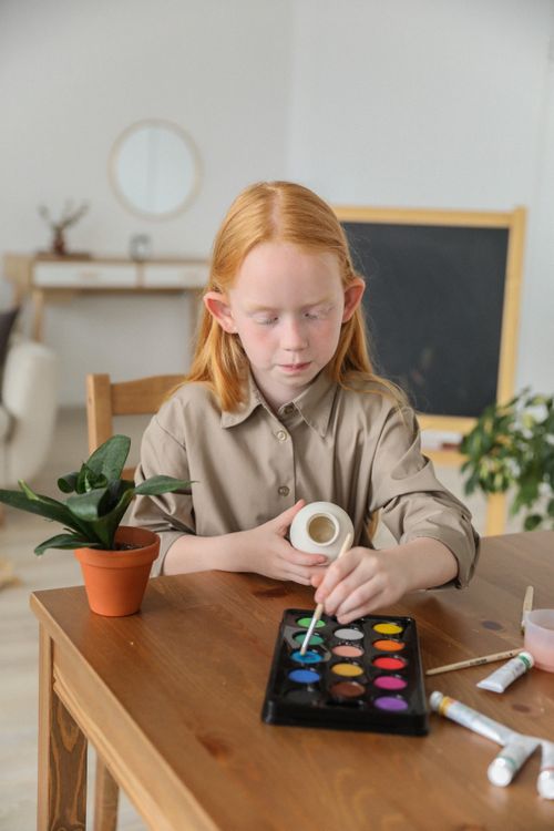 Free Concentrated girl siting at wooden table and painting small ceramic vase at home Stock Photo