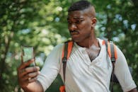 一个阴郁的非洲裔美国人背着包，在阳光明媚的日子里，举着智能手机对着绿树看屏幕