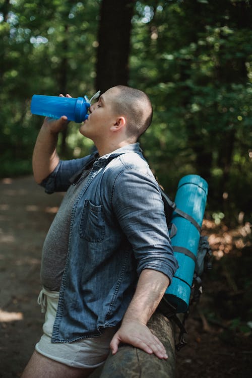 Free Side view of man in casual clothes with backpack drinking bottle of water while resting in park during hike in daytime Stock Photo