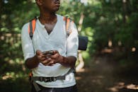 Crop unrecognizable African American man with backpack and smartphone searching route while going astray in green forest