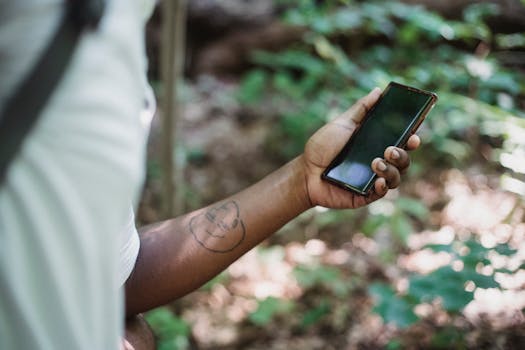 Crop unrecognizable African American man with tattoo standing in green forest and browsing mobile phone