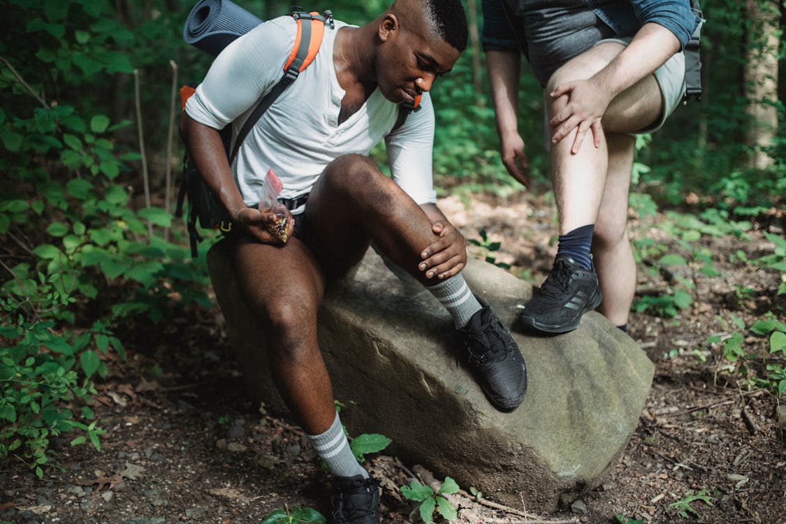 Free Multiethnic men in sportswear with backpacks resting together and searching for ticks on legs in green lush forest during trek Stock Photo
