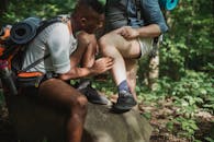 Multiracial men in activewear with backpacks resting on stone and checking ticks while having break during trekking