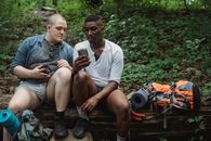 Full body of concentrated multiracial male hikers surfing internet while resting on old brown horizontal log in forest in daytime