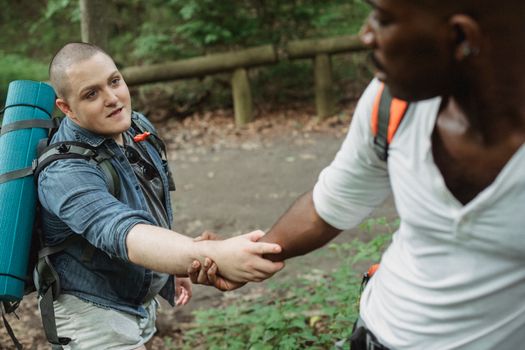 Unrecognizable black adventurer providing hand for happy plump male friend with big backpack and rolled rug in woodland in daytime