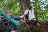Focused African American traveler lending hand for faceless backpacker next to tree in forest in daytime