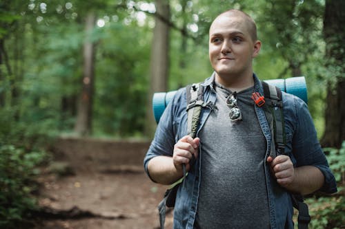 Smiling plus size male backpacker standing on path in green dense forest during hiking