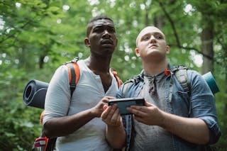 From below of diverse lost travelers with backpacks exploring woodland and searching way in cellphone