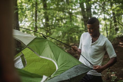 Free Cheerful African American male in casual clothes putting tent in green lush forest during vacation Stock Photo