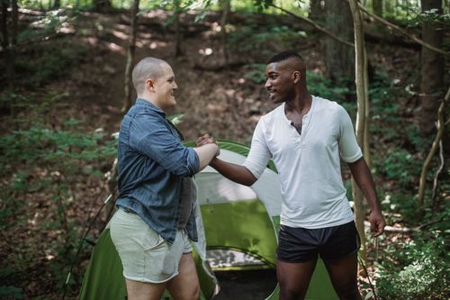 Free Black male tourist enjoying vacation with friend in summer forest while standing and shaking hands near green tent Stock Photo