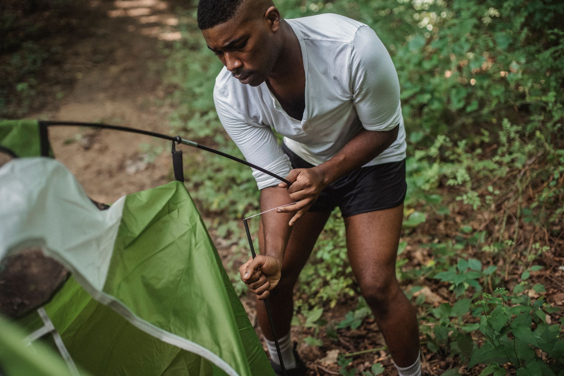 From above of young African American guy in shorts and white shirt standing and setting up green tent in forest on sunny summer day