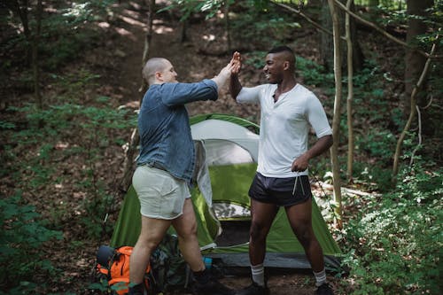 Young cheerful guy in jeans shirt and shorts giving high five to African American friend traveler during vacation with tent in woodland