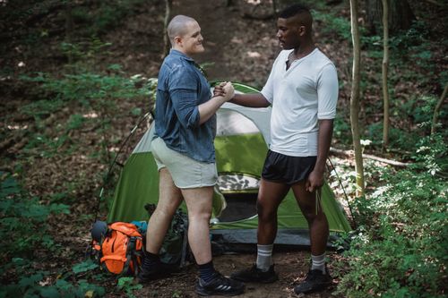Free Positive black guy shaking hands with friend in front of camping tent among green trees Stock Photo