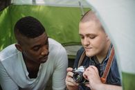 Young male showing sharing retro photo camera with African American friend while sitting in green tent