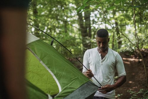 Free Serious young African American guy in white shirt inserting stick of tent in hole during hiking in summer forest Stock Photo
